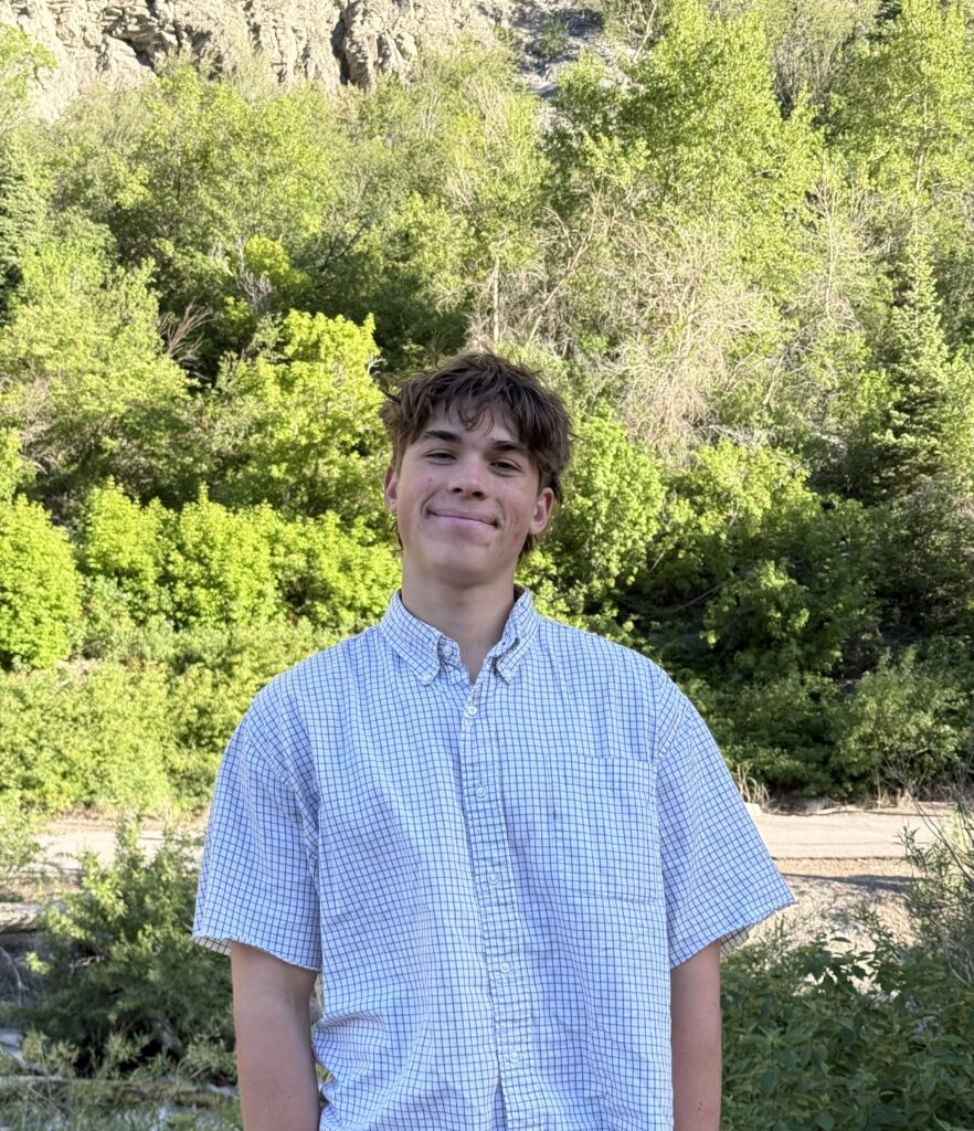 Boy standing in front of a tree, with a white shirt, and brown hair.