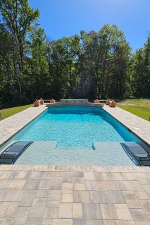 Modern rectangular pool with sun shelf loungers, raised tile water wall, and stone paver deck surrounded by trees.