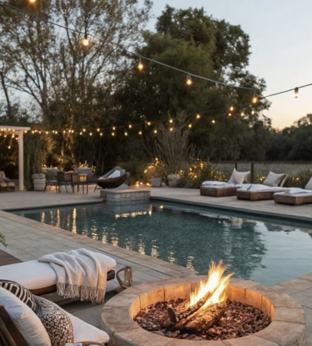 Poolside fire pit with cozy lounge seating, string lights, and a modern pool in a landscaped backyard at sunset